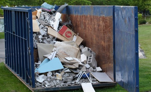A large metal skip container painted in dark blue, positioned outdoors on a grassy area, filled with a mixture of construction and domestic waste including broken concrete slabs, plasterboard, cardboard boxes, and various pieces of damaged drywall and wood. The interior of the skip appears rusted from prolonged exposure to the elements, with the waste piled unevenly, spilling over slightly at the top. The surrounding environment features a well-maintained lawn and some trees in the background, suggesting a suburban or semi-rural setting. This scene, associated with rubbish removal services such as those provided by Commercial Waste Camberley, illustrates the collection of bulky waste materials typical of local waste management in the Camberley area. The image emphasizes the importance of proper disposal of mixed waste and the role of professional rubbish removal providers in managing substantial refuse loads effectively.