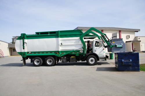 A large commercial rubbish collection vehicle, equipped with a green and white body and a hydraulic lifting arm, is parked on a paved driveway in front of a modern residential building. The vehicle's body appears to be made of metal with a smooth finish, and it is positioned centrally in the image, with the hydraulic arm extended slightly over a dark blue communal waste bin placed nearby. Another similar blue bin is situated to the left of the first, both on the right side of the vehicle. The background features part of the residential building with light exterior walls, multiple windows, and a flat roof, indicating a contemporary urban setting. The environment is well-lit with natural daylight, with no other vehicles or objects visible, conveying an organized waste collection scene typical of commercial waste services in Camberley.