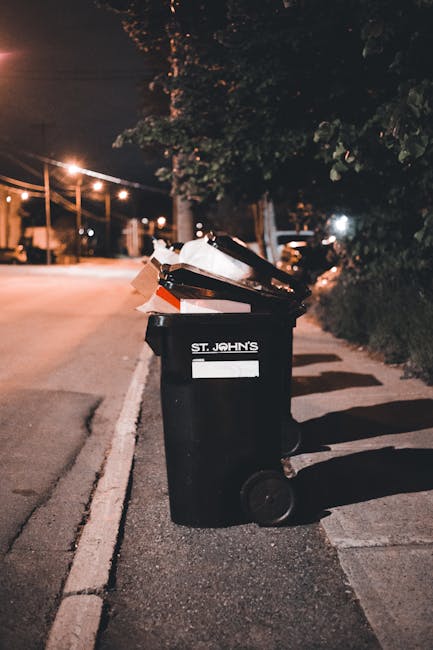 A black residential waste bin placed on the pavement beside a curbside street at night, with its lid open revealing assorted rubbish including paper, cardboard, and plastic items. The bin is marked wi