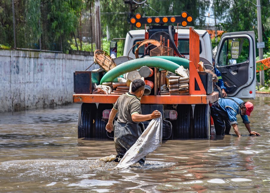 Two workers are seen in a flooded outdoor area, engaged in rubbish removal and cleanup activities. One worker, standing in shallow water, is holding a white plastic bag and appears to be preparing for